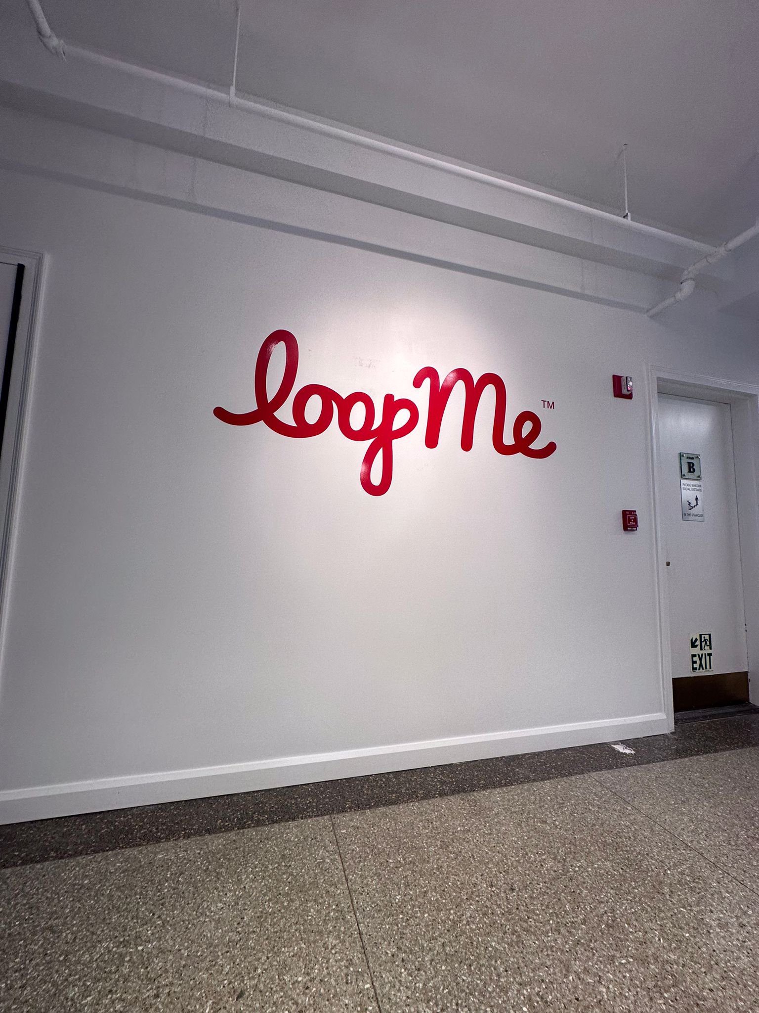 A modern office hallway featuring a large, red logo for LoopMe. The logo is displayed in a stylized font and includes the trademark symbol. An exit sign is visible on the right side of the image.